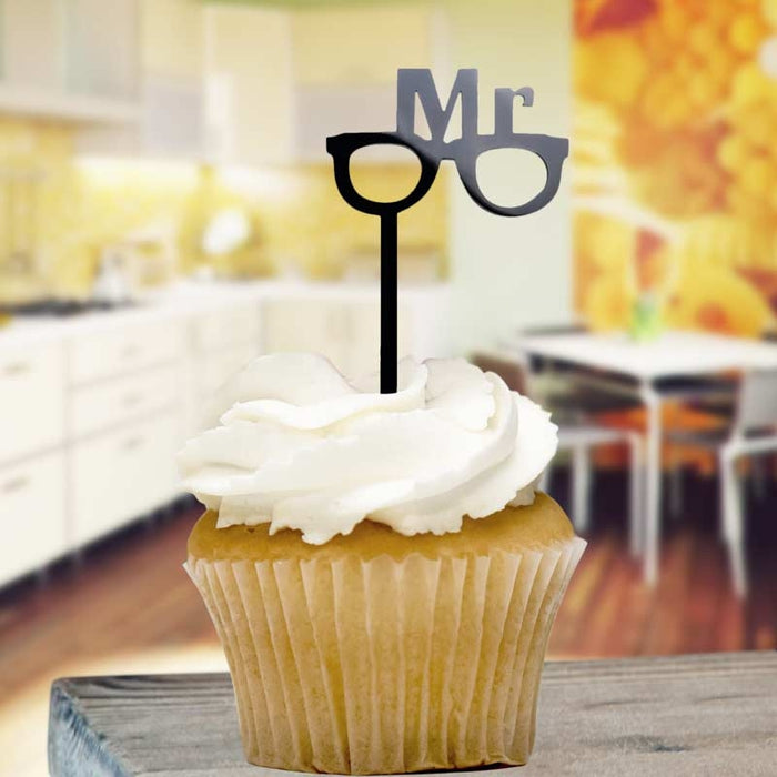 Cupcake with white frosting and a 'Mr' sign on top, set against a blurred indoor background.