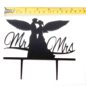 Decorative cake topper with 'Mr' and 'Mrs' text, featuring a couple with wings, on a white background.